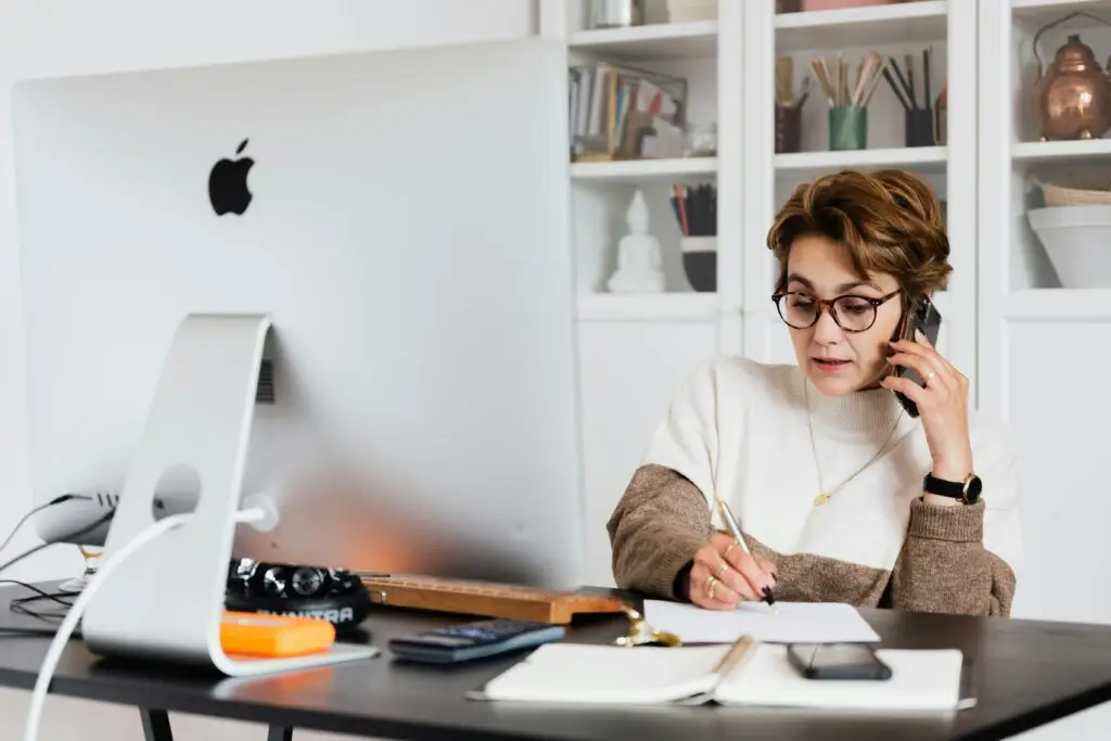 A businesswoman multitasks on the phone while writing notes at her home office desk, showcasing modern productivity.